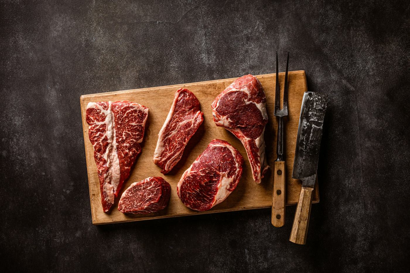 Assorted raw marbled beef steaks on a wooden board with a carving fork and cleaver