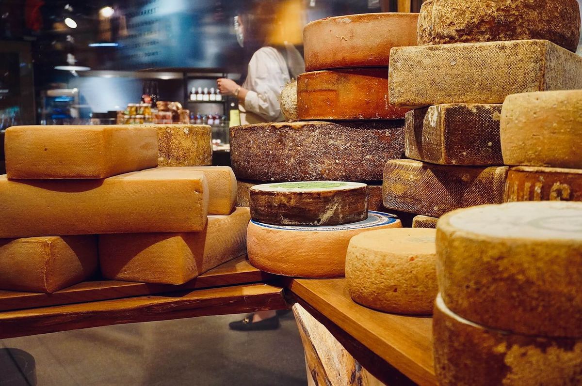 Stacks of whole aged cheese wheels and blocks with thick natural rinds at an artisan cheese counter, with a cheesemonger working behind the display.